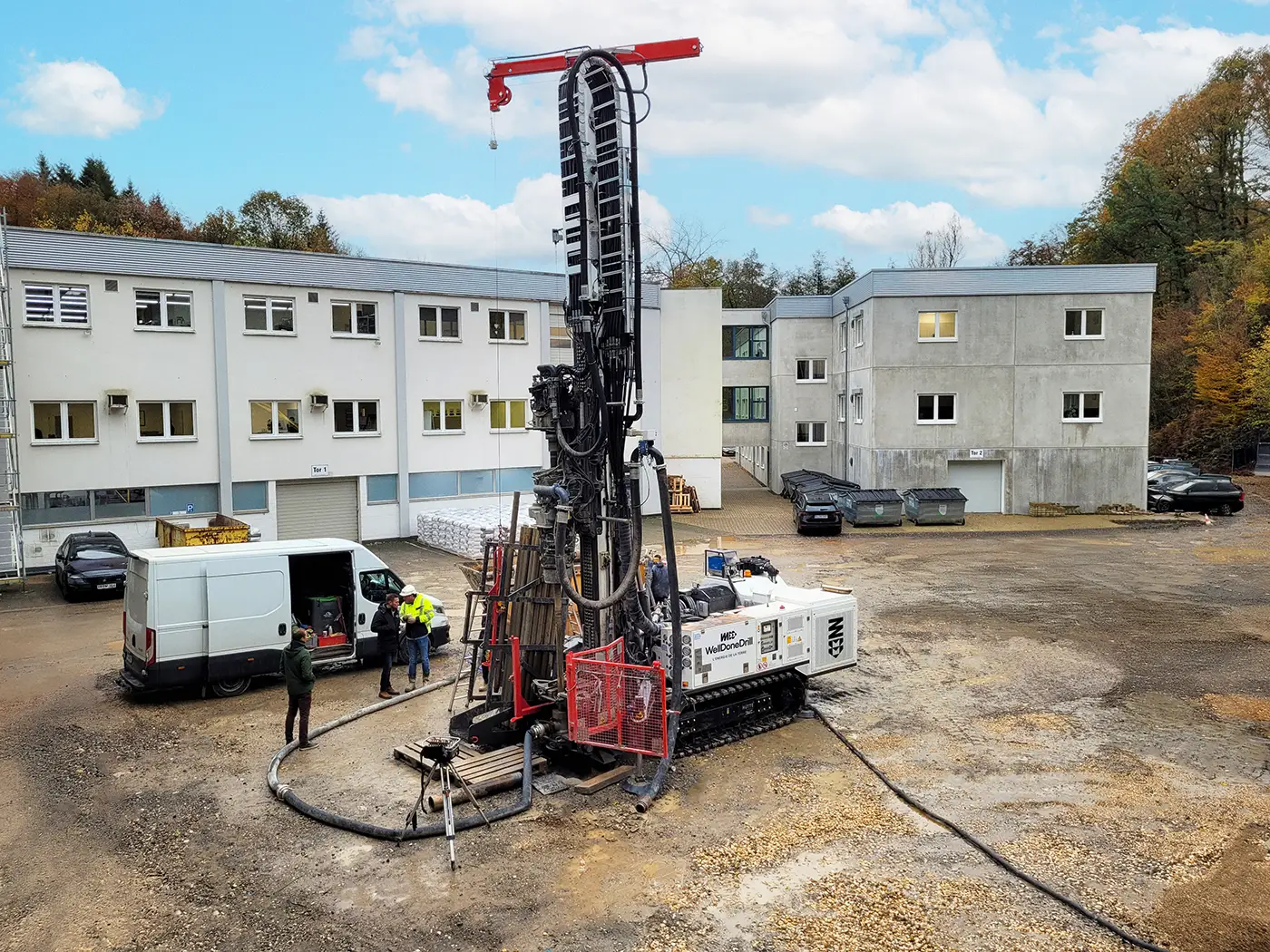 wdd-machine-forage Machine de forage géothermique en action sur un chantier en Belgique, avec des techniciens travaillant à côté de véhicules et d'un bâtiment administratif.